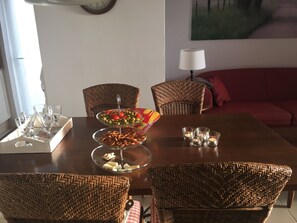 Un comedor con sillas de mimbre, una mesa de comedor con un centro de mesa de cristal y un sofá rojo al fondo.