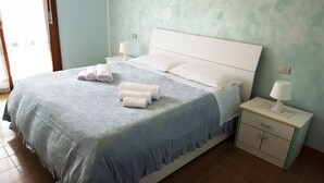 A neatly made bed with a blue bedspread and white pillows, two rolled towels on the bed, a white nightstand with a lamp, and a small cabinet with a green handle.