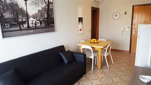 A room with a black sofa, a wooden table with chairs, a fruit bowl, a calendar, and a wall-mounted clock.