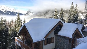 Une cabane en bois recouverte de neige, dotée d’un balcon, entourée d’arbres chargés de neige et avec des montagnes en arrière-plan.