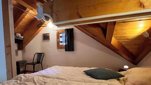 Une chambre mansardée chaleureuse avec un lit superposé, des poutres en bois, une petite fenêtre avec des rideaux, une chaise et une table de chevet avec une lampe.