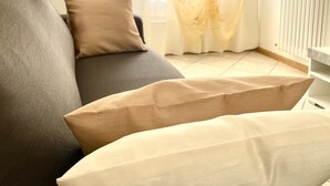 Una sala de estar con un sofá gris, cojines color beige, un radiador blanco y cortinas traslúcidas con estampado floral.