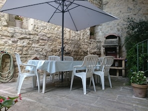 Een terras met stenen muren, een tafel en stoelen onder een grote parasol, omringd door plantenpotten en een barbecue.