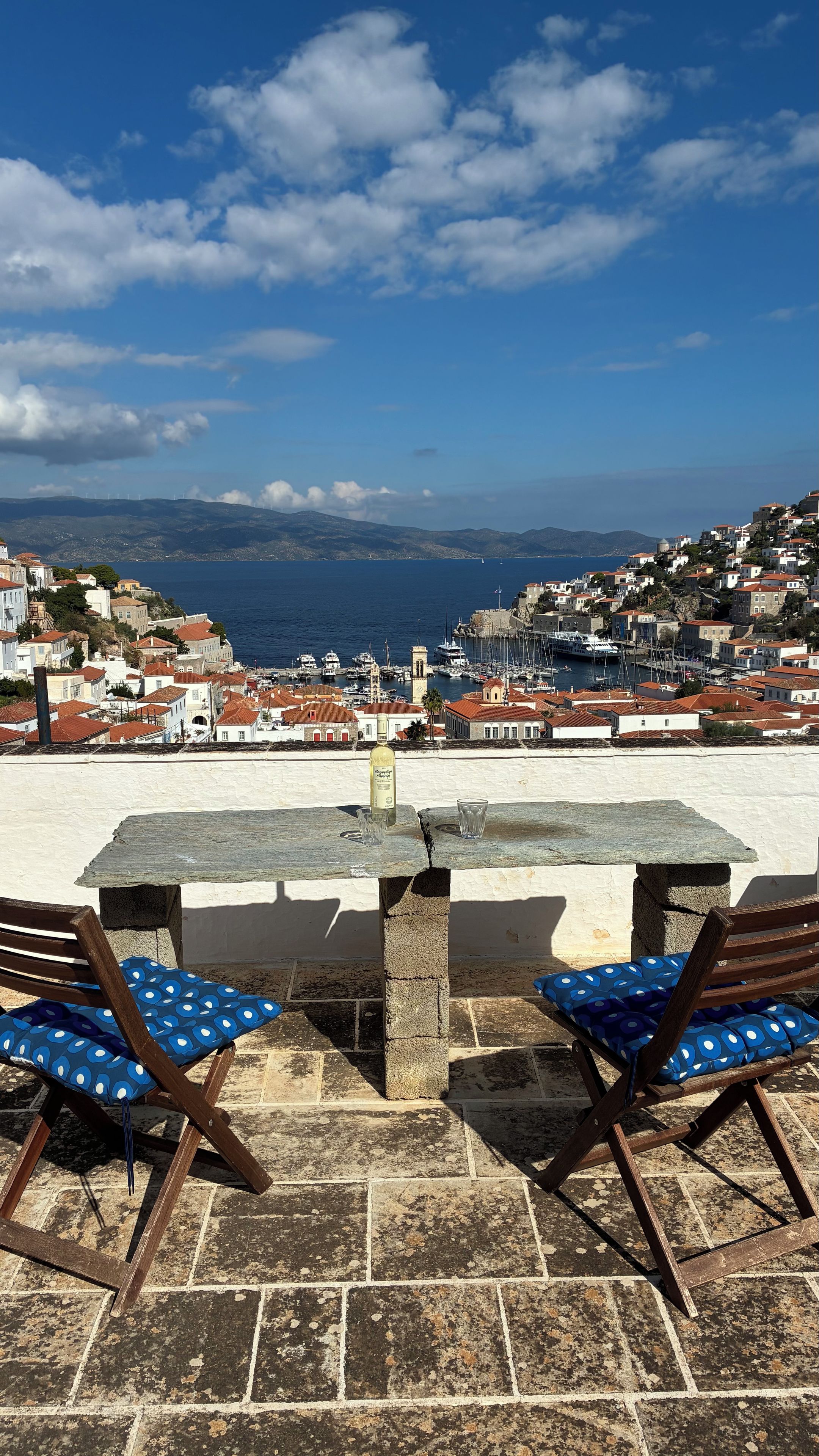 A seaside terrace with a stone table and two wooden chairs with blue cushions, overlooking a coastal town with buildings and boats.