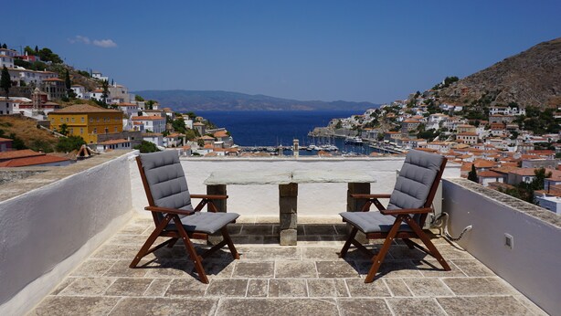 Una terrazza con due sedie in legno e un tavolo, con vista su una località costiera caratterizzata da edifici e un mare cristallino.