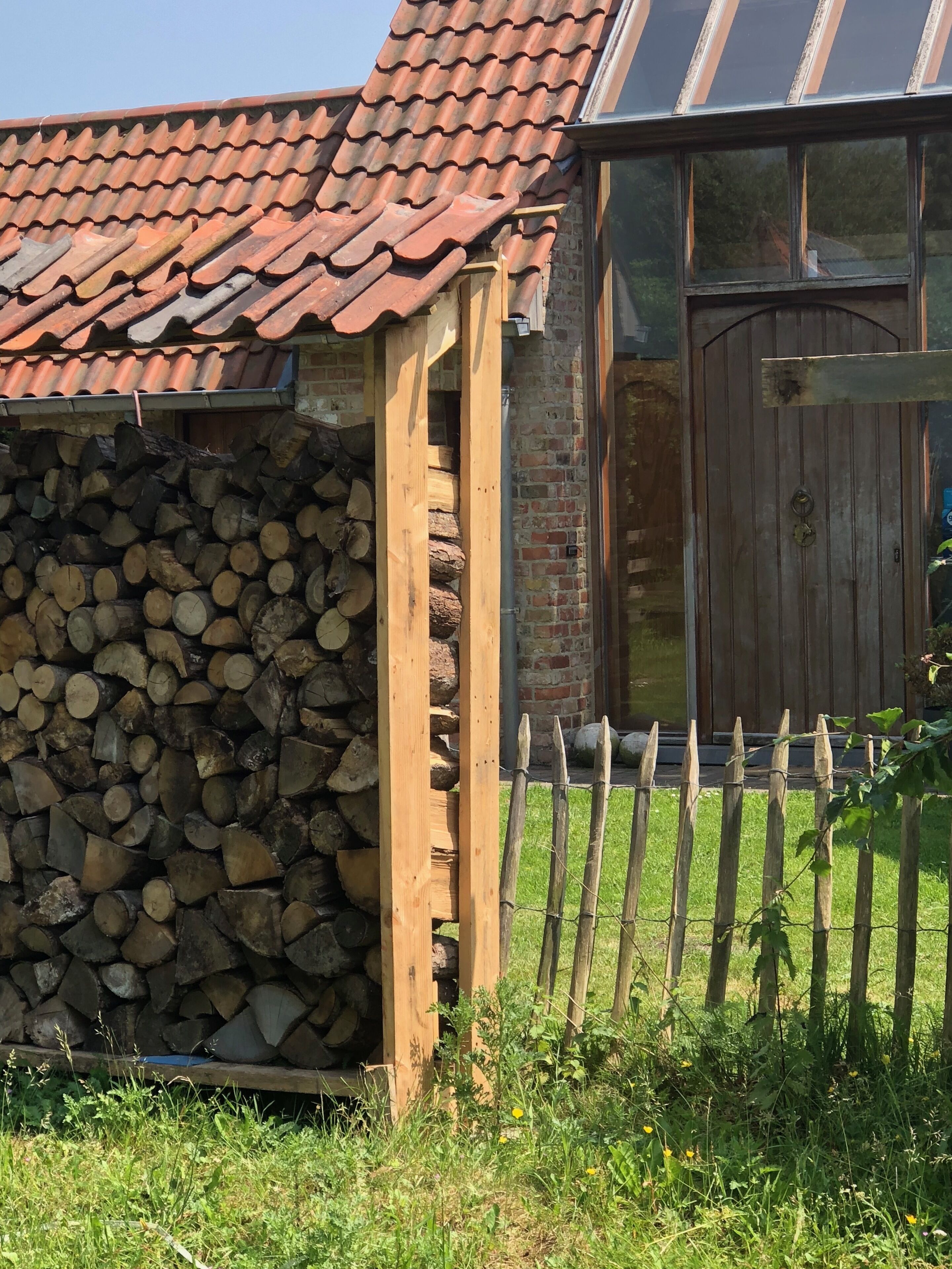 Una estructura rústica de madera con techo de tejas, una pila de leña y una valla de madera.