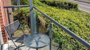 En balkon med et glasbord og stole, der ud til en vej og et vandområde.