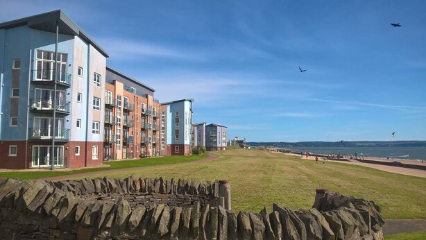 En række høje boligbygninger med altaner, en grøn græsplæne foran og en strand i baggrunden.