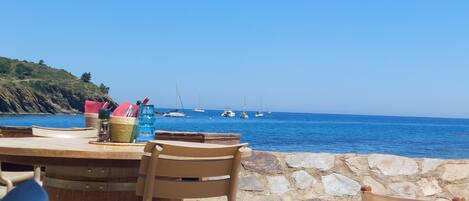 A table with chairs and a stone wall, overlooking a calm sea with boats.