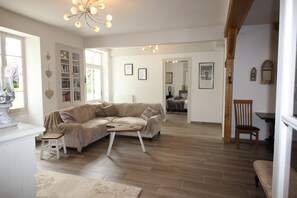 A spacious living room with a beige sofa, a wooden coffee table, and a white armchair. There is a bookshelf with books and decorative items, and a chandelier hanging from the ceiling.
