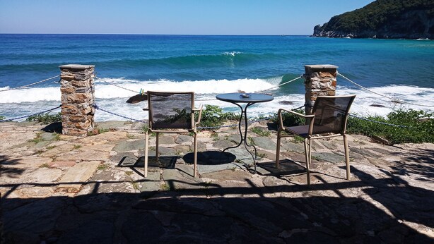 Un patio de piedra con dos sillas y una pequeña mesa, con vistas a una playa donde las olas rompen en la orilla.