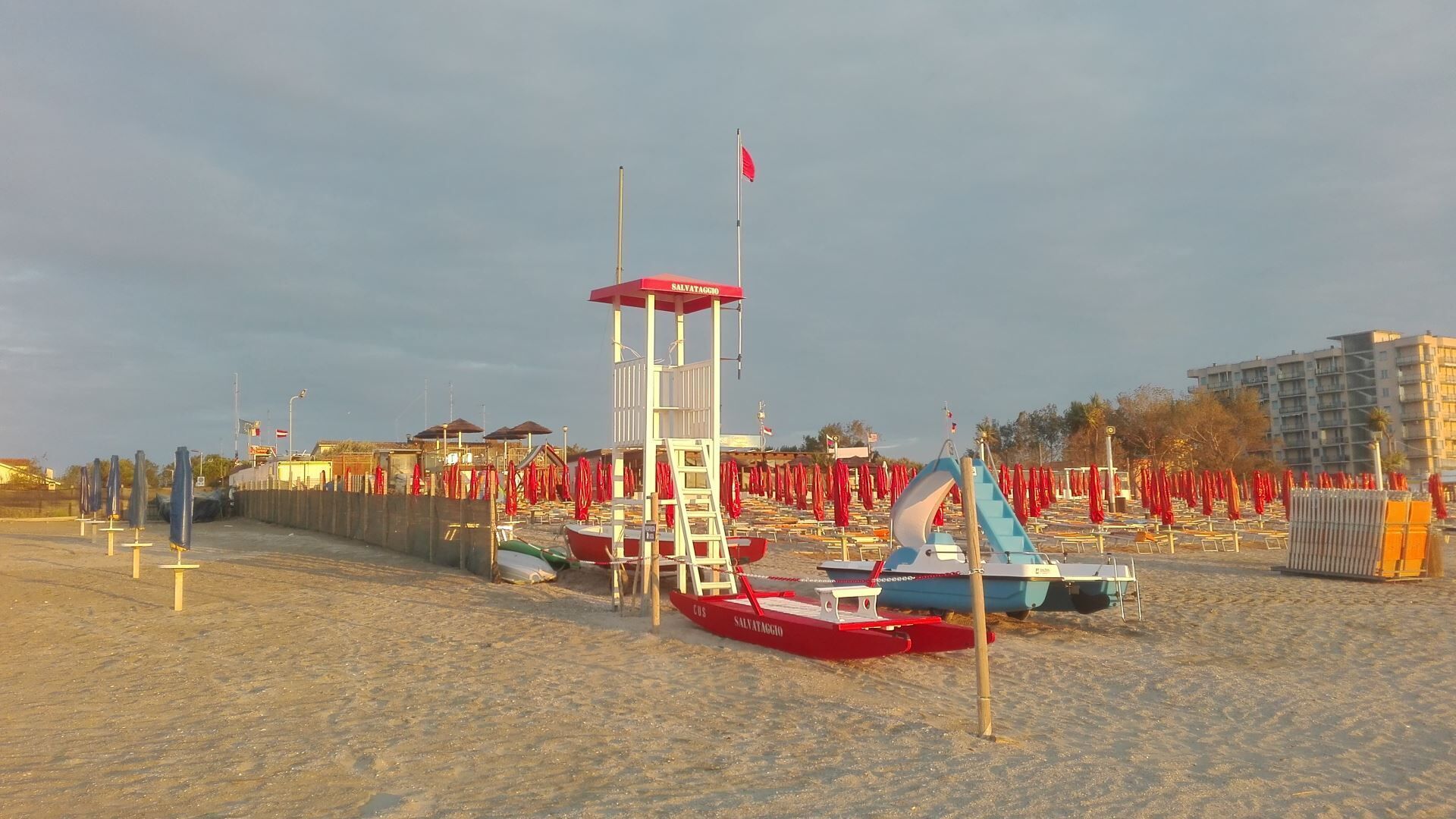 Una torre de salvavidas con una bandera, sombrillas de playa y una silla para salvavidas en una playa arenosa.