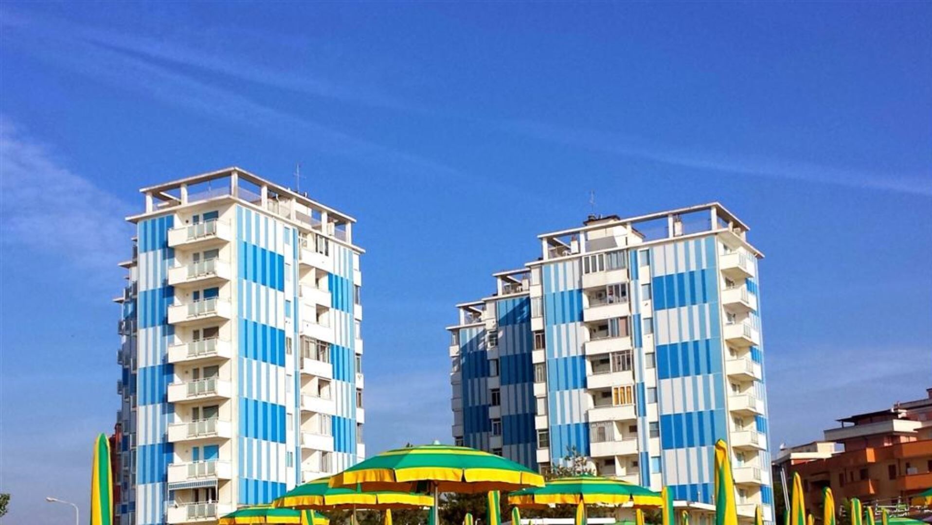Two tall, multi-story buildings with balconies, likely residential or hotel units, under a clear blue sky.
