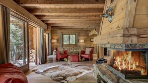 Ein rustikales Blockhaus-Interieur mit Kamin aus Stein, Holzbalkendecke, Essbereich mit roten Stühlen und einer großen Glasdoor, die nach draußen führt.