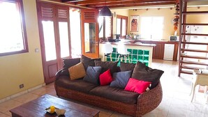 Een woonkamer met een bruine rieten bank, een houten salontafel met boeken, en een keuken in de achtergrond.