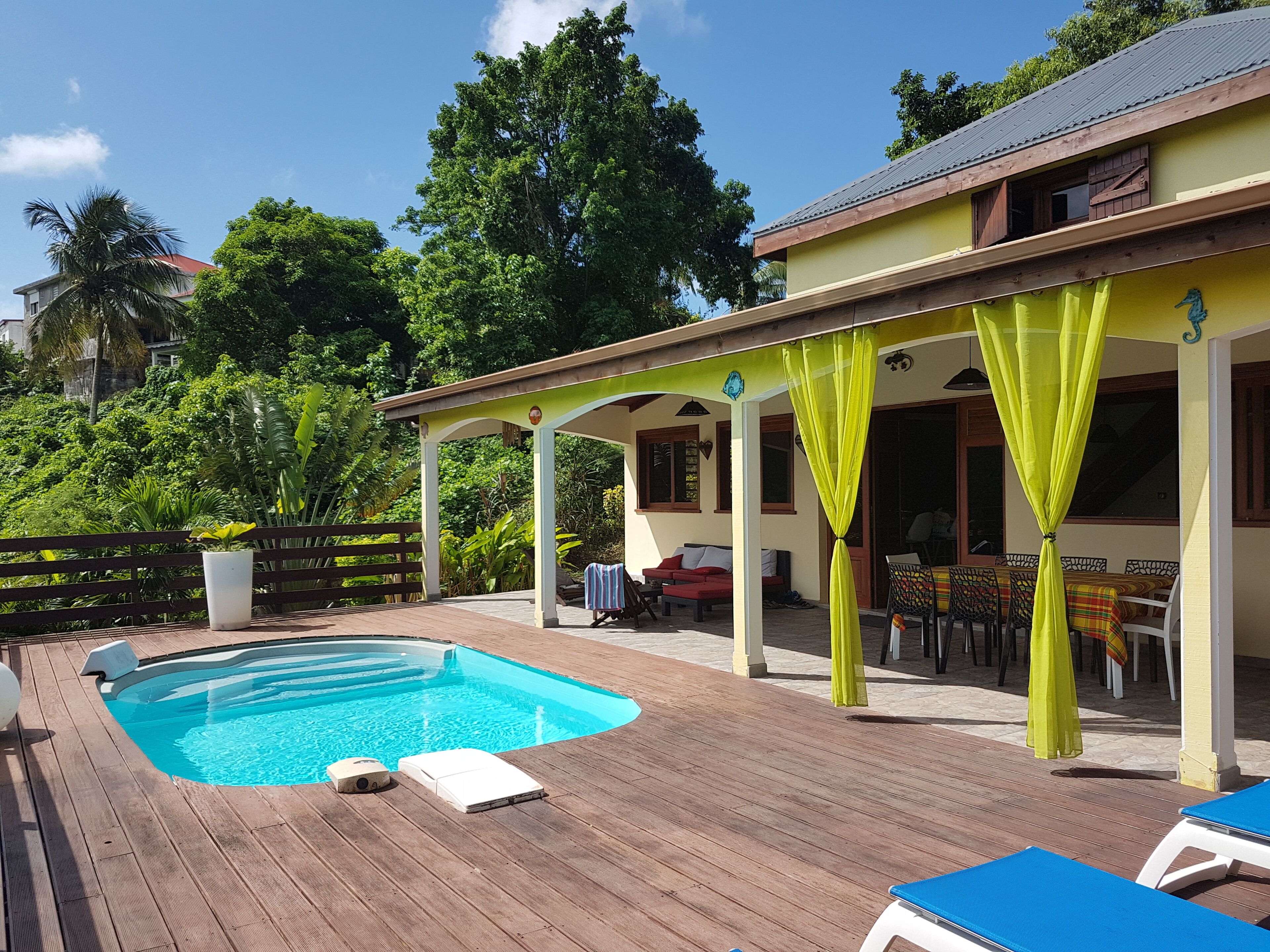 Ein Poolbereich mit Holzterrasse, überdachtem Sitzbereich und einem Gebäude mit gelber Fassade.