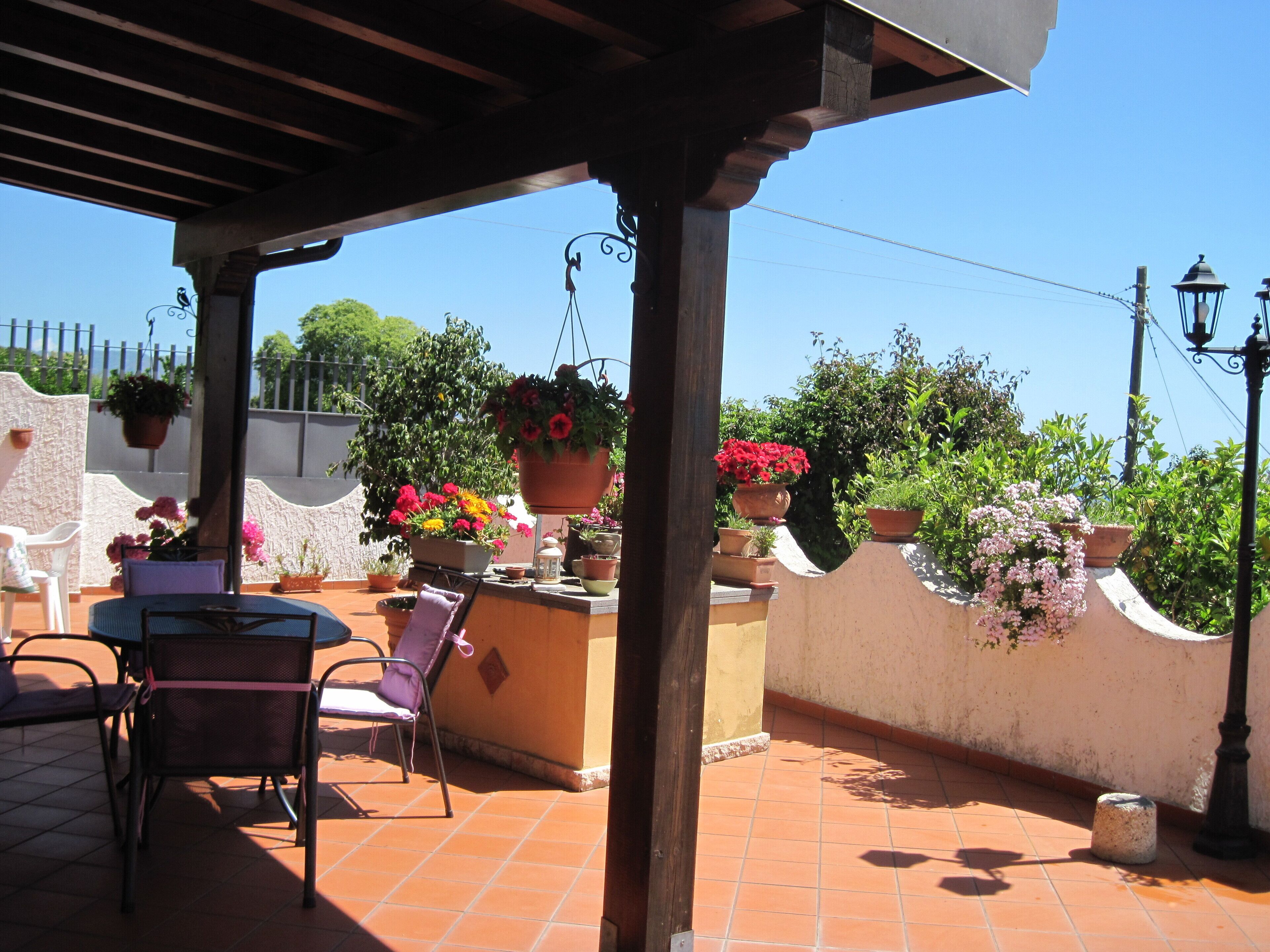 A covered patio area with a table and chairs, surrounded by potted plants and a white wall.