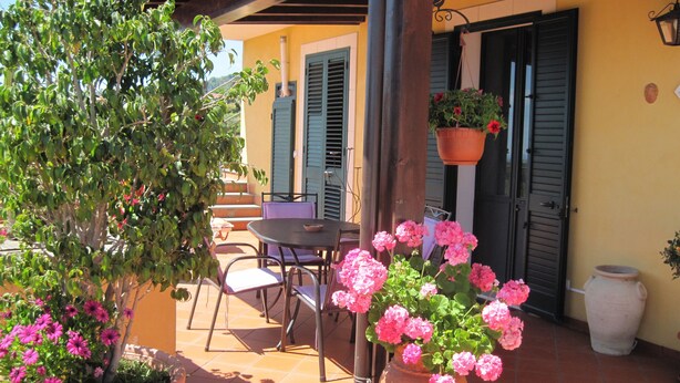 Un patio al aire libre con una mesa y sillas, una maceta colgante y un jarrón grande con flores rosadas.