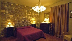 Un dormitorio con pared de piedra, una araña, una cama con manta roja, dos mesitas de noche con lámparas y una silla.