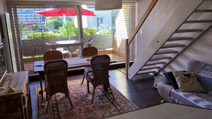 Una sala de estar moderna con una escalera, un comedor con sillas de mimbre, un sofá con cojines estampados y vistas al exterior con vegetación.