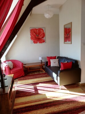 A cozy living room with a red armchair, a sofa with red and white cushions, a wooden coffee table, and a striped rug. There are two large red flower paintings on the walls.