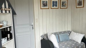 Una habitación acogedora con una puerta blanca, un sofá negro con cojines estampados, una estantería con electrodomésticos y cuadros en la pared.