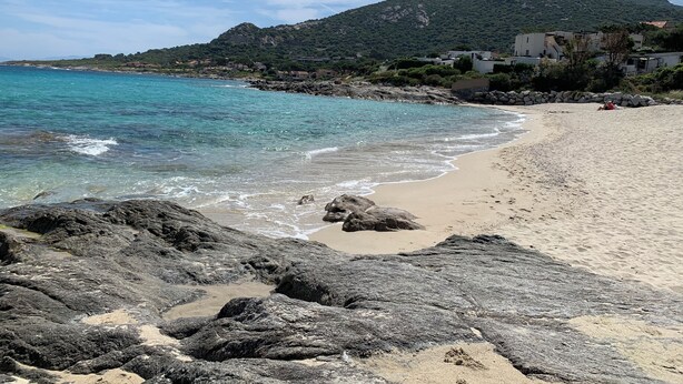 A beach with rocky foreground, clear turquoise waters, and a mountain in the background.