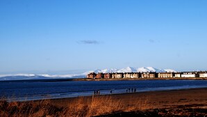 Vista costera con edificios, una playa y montañas nevadas al fondo.