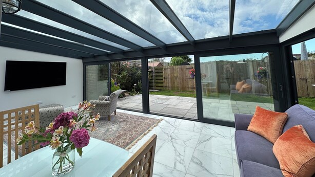 Un patio moderno con techo de vidrio, televisor de pantalla plana, mesa de comedor con un jarrón de flores y una zona de estar con cojines.