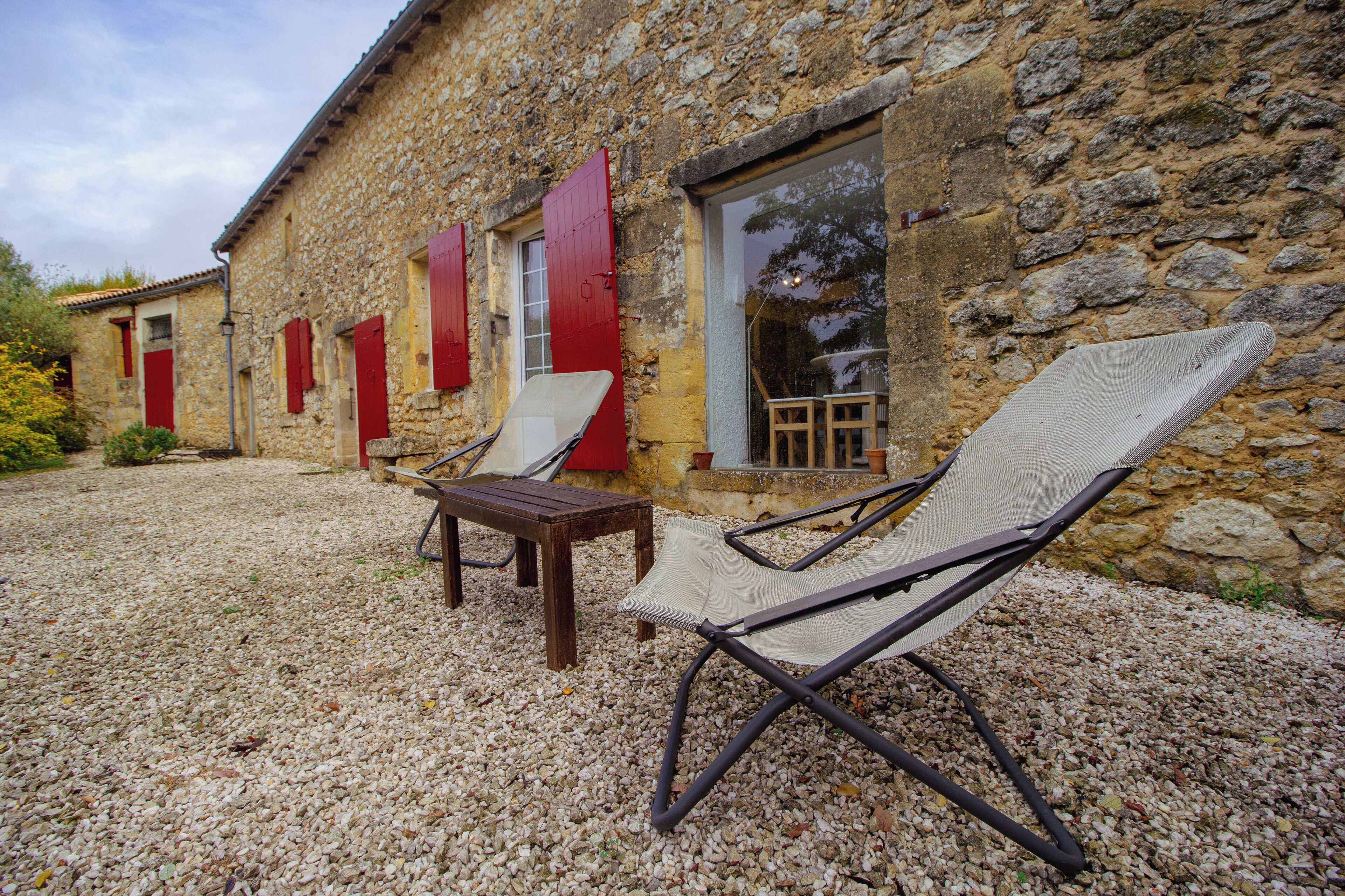 A stone building with red shutters, two lounge chairs, and a wooden table.