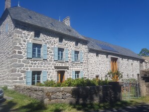 A stone building with multiple windows and shutters, a sloped roof, and a small garden area.