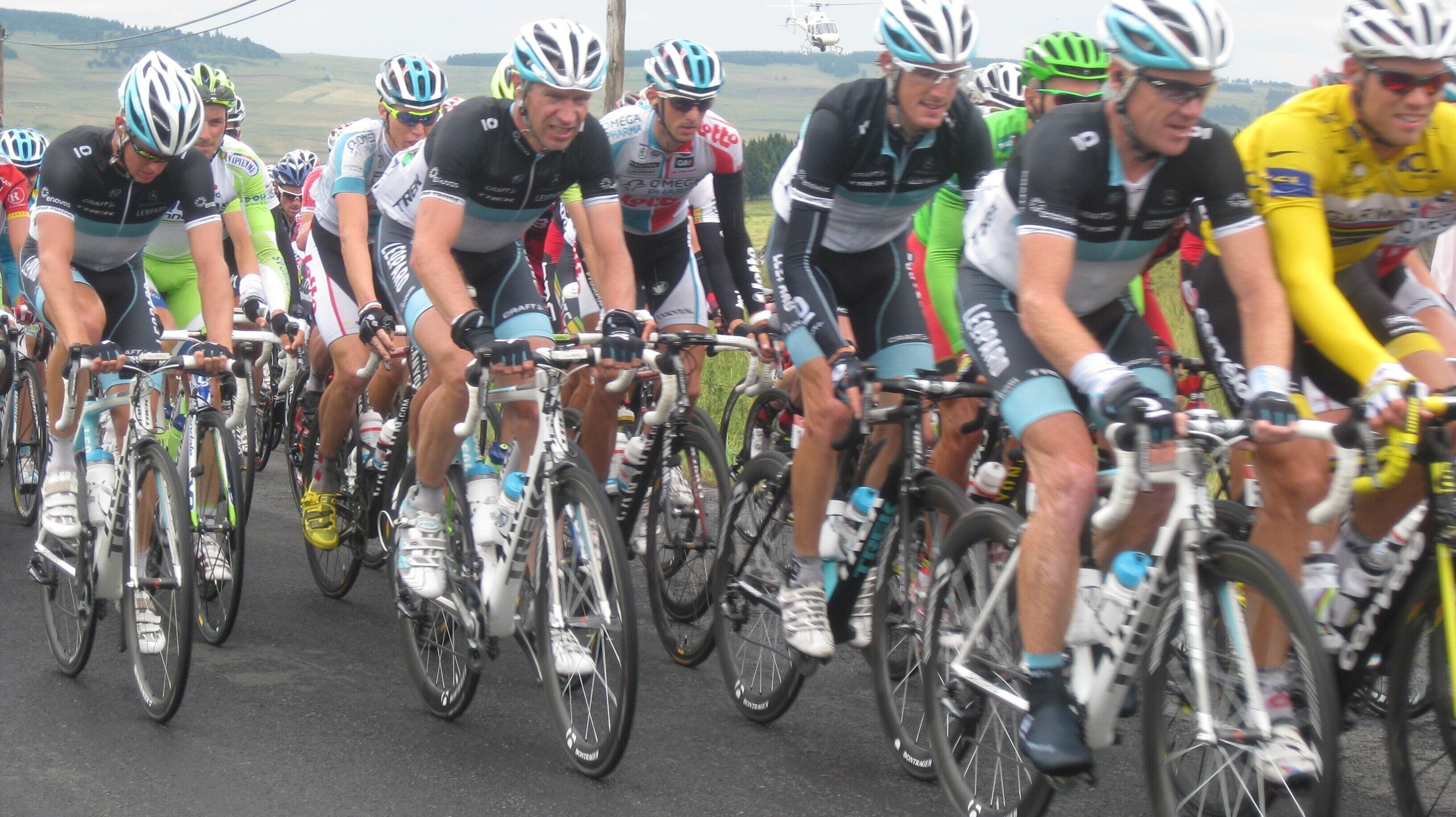 Un groupe de cyclistes en mouvement, portant différents maillots d’équipe et des casques, sur une route asphaltée.
