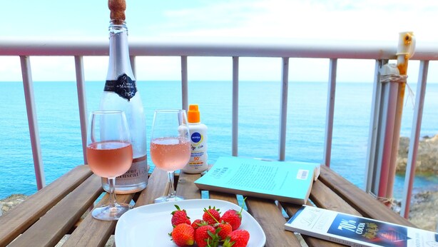 A wooden table with a bottle of rosé wine, two glasses of rosé wine, a bottle of sunscreen, a plate of strawberries, and a book on a balcony overlooking the sea. - Collioure Seafront Apartment with Parking - Falco (Collioure)