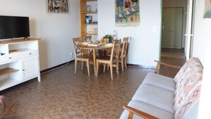 A living room with a TV on a stand, a dining table with chairs, a sofa, and a large tapestry on the wall. - Collioure Seafront Apartment with Parking - Falco (Collioure)