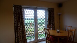A room with a dining table and chairs, a window with curtains, and a view of a green landscape. - Welcome to Tuolumne Cottage (Denbigh)