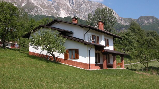 A two-story house with a sloped roof, brick chimney, and a small porch with a bench. It is surrounded by a grassy hill and trees, with mountains in the background.