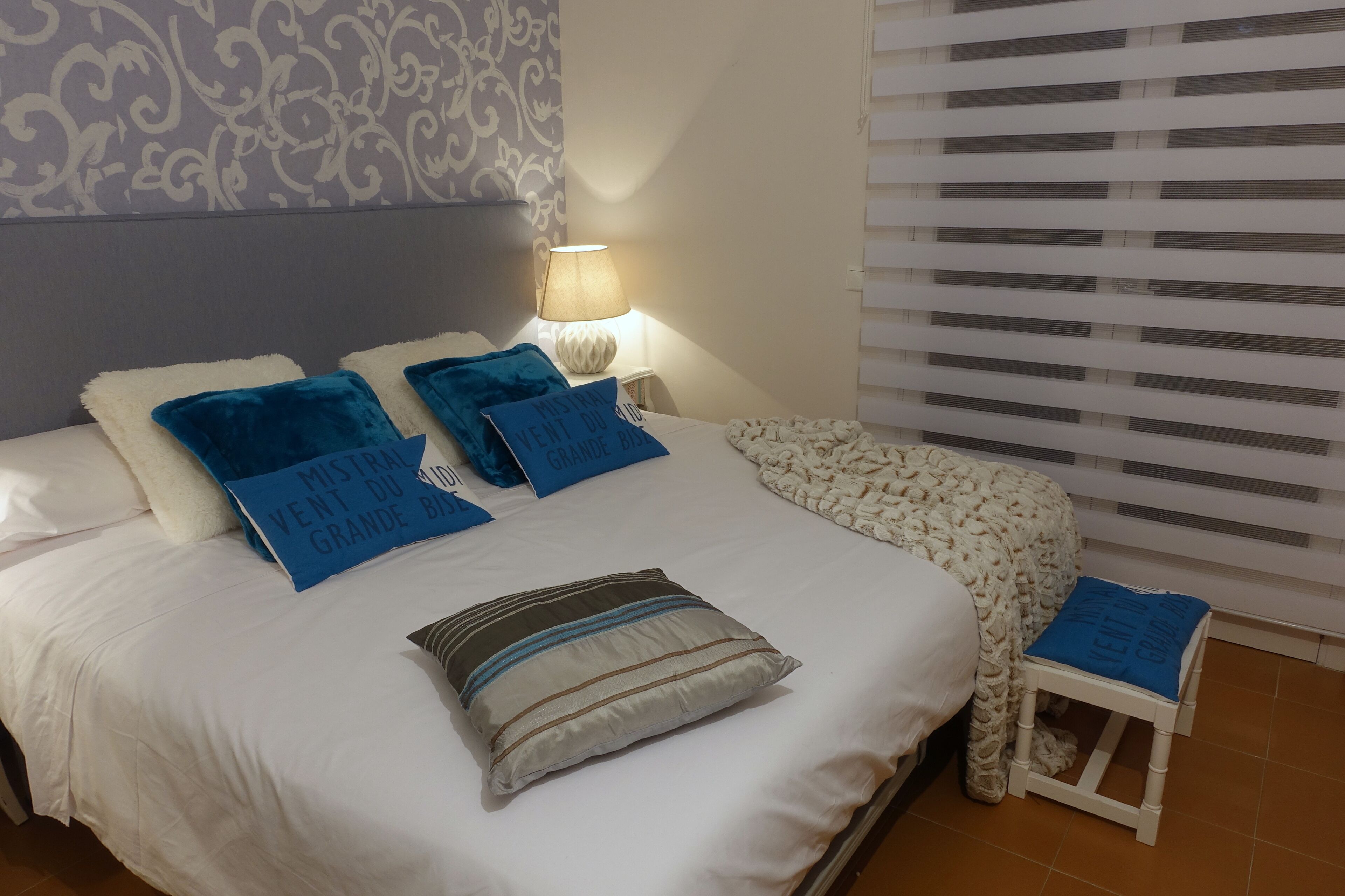 A neatly made bed with multiple pillows, a bedside lamp, and a small stool.