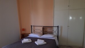 A neatly made bed with white pillows and folded towels, a wooden nightstand with a red lamp, and a wardrobe with a door lock. - Katerina Apartments ,( 2 ) One-Bedroom Apartment with Balcony (Kalamata)