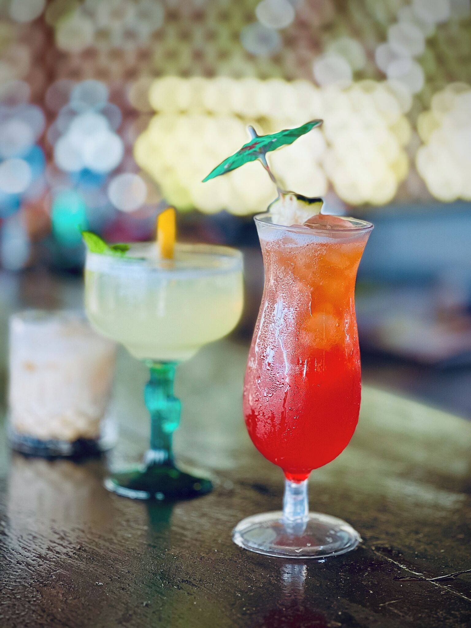 Two cocktails on a wooden table with a blurred background of lights.