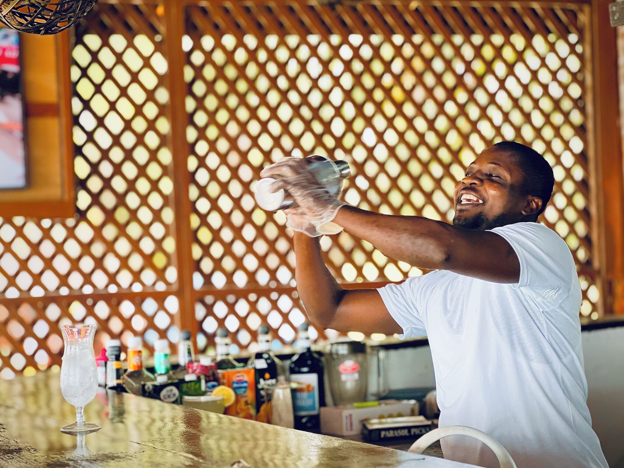 A person pouring a drink into a glass at a bar with a lattice backdrop.