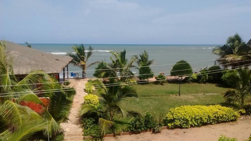 A coastal view with a pathway leading to a beach, surrounded by palm trees and a thatched-roof structure.