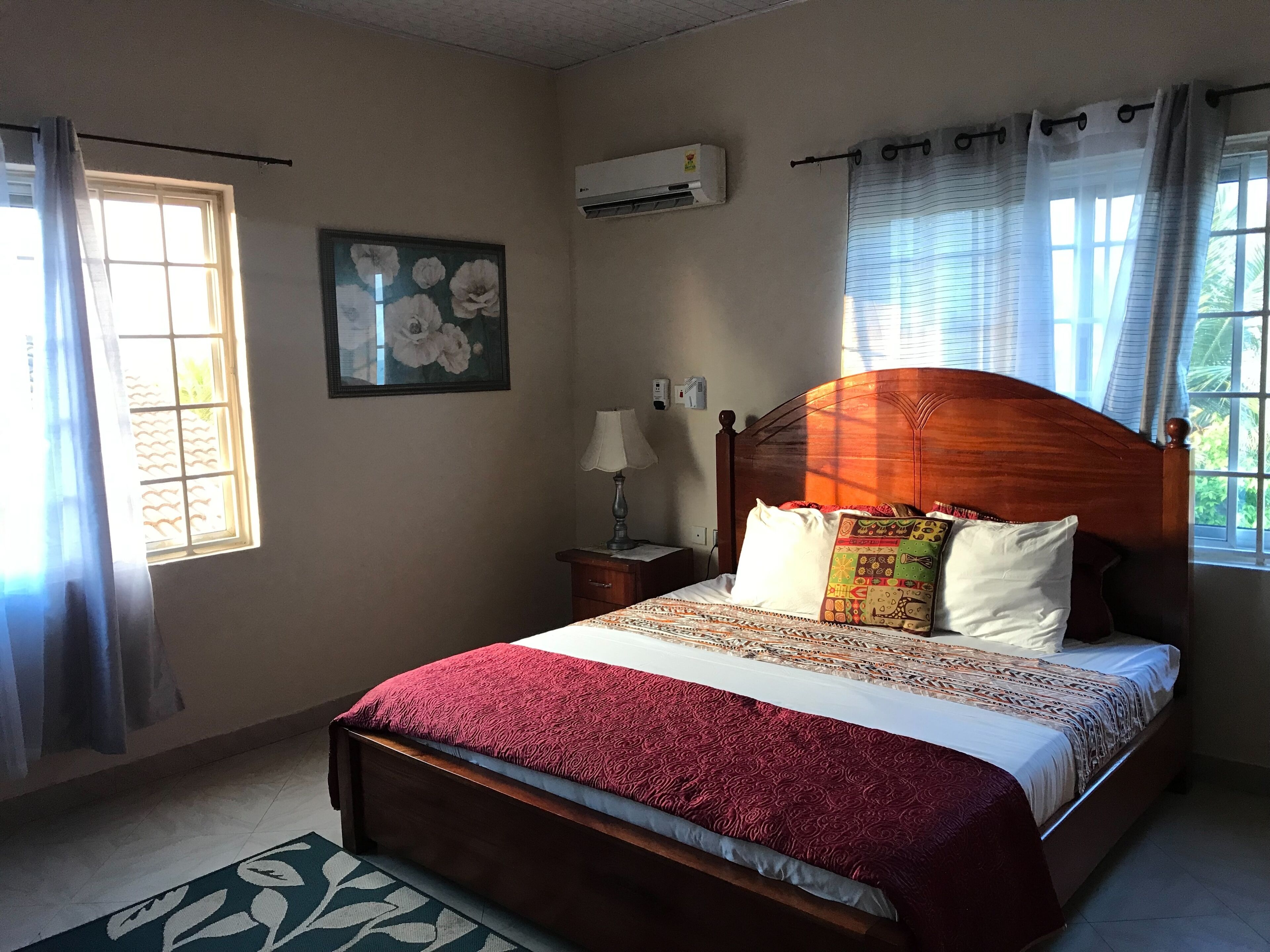 Un dormitorio con cama de madera, una mesita de noche, una lámpara, aire acondicionado instalado en la pared, una ventana con cortinas y un cuadro con motivos florales.