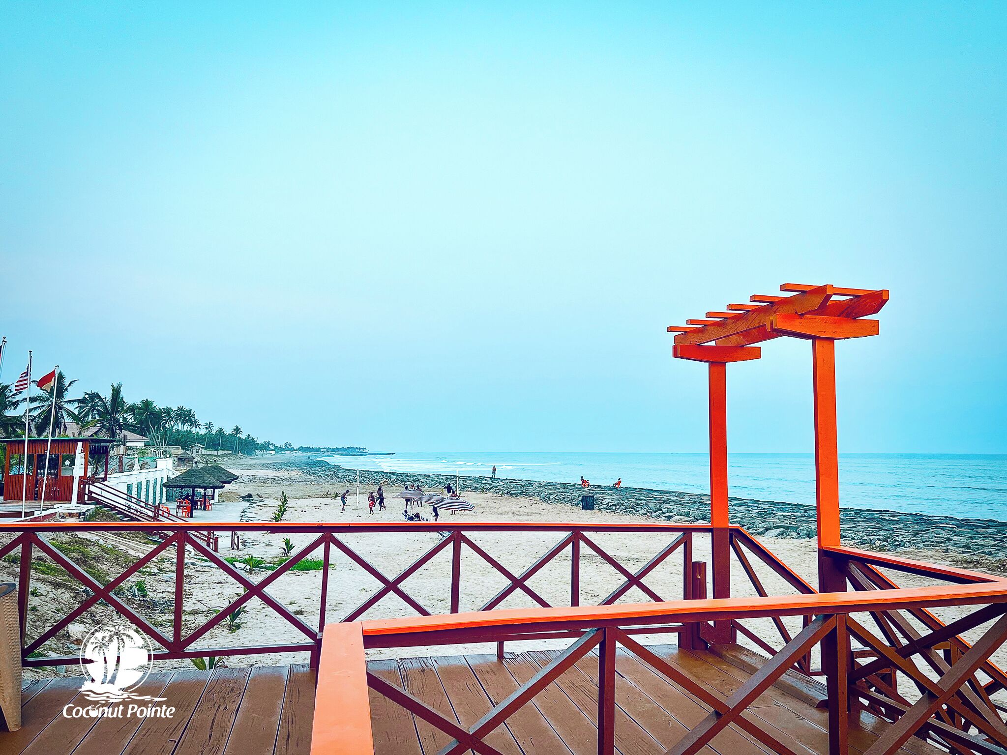 Vista a la playa desde una terraza de madera con barandilla roja, palmeras y cielo despejado.