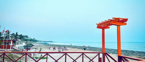 Vista a la playa desde una terraza de madera con barandilla roja, palmeras y cielo despejado.