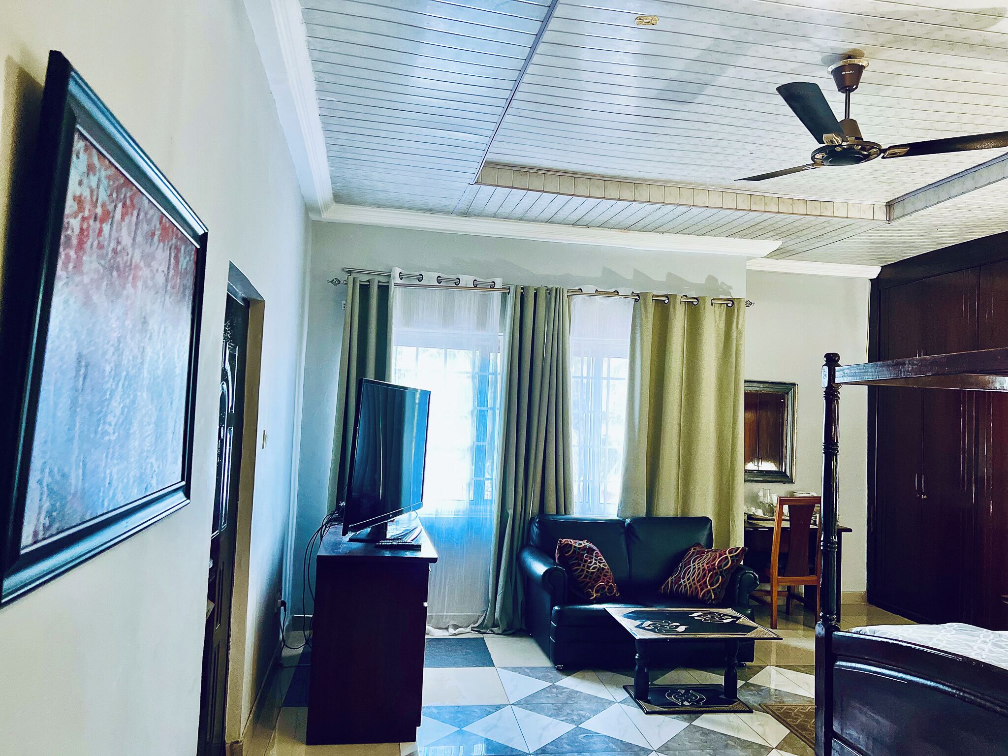 Une chambre avec un lit, un bureau, une télévision, un ventilateur de plafond et une fenêtre avec des rideaux.