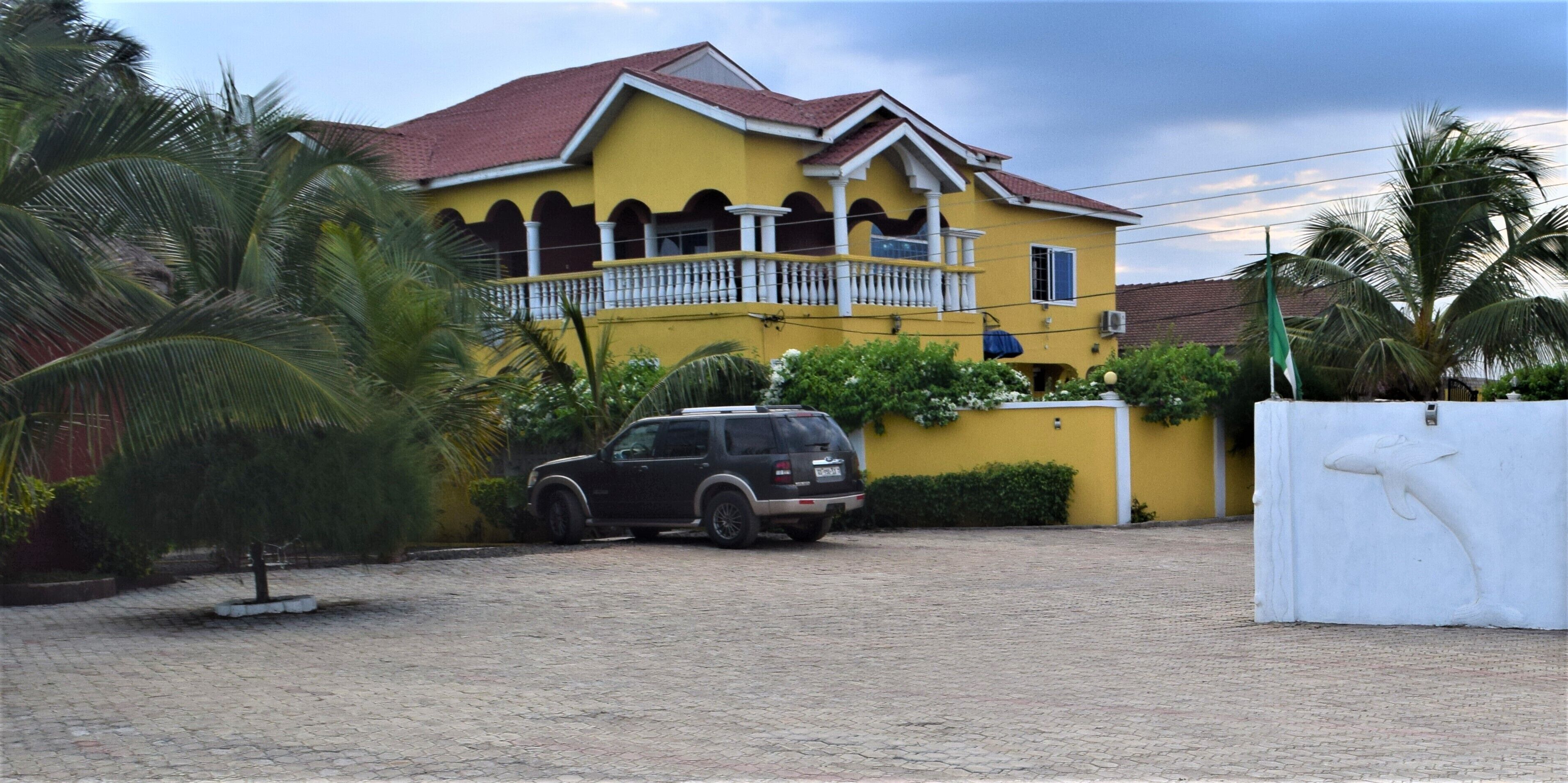 A yellow house with a balcony, a parked SUV, and a white fence with a dolphin sculpture.
