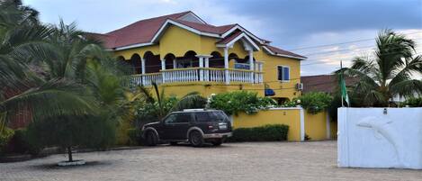 A yellow house with a balcony, a parked SUV, and a white fence with a dolphin sculpture.