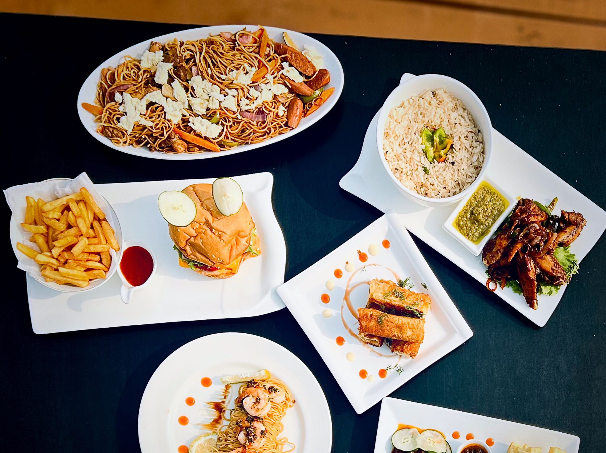 Une variété de plats, notamment des nouilles, du riz, un hamburger, des frites et du poulet grillé, le tout servi dans des assiettes blanches.