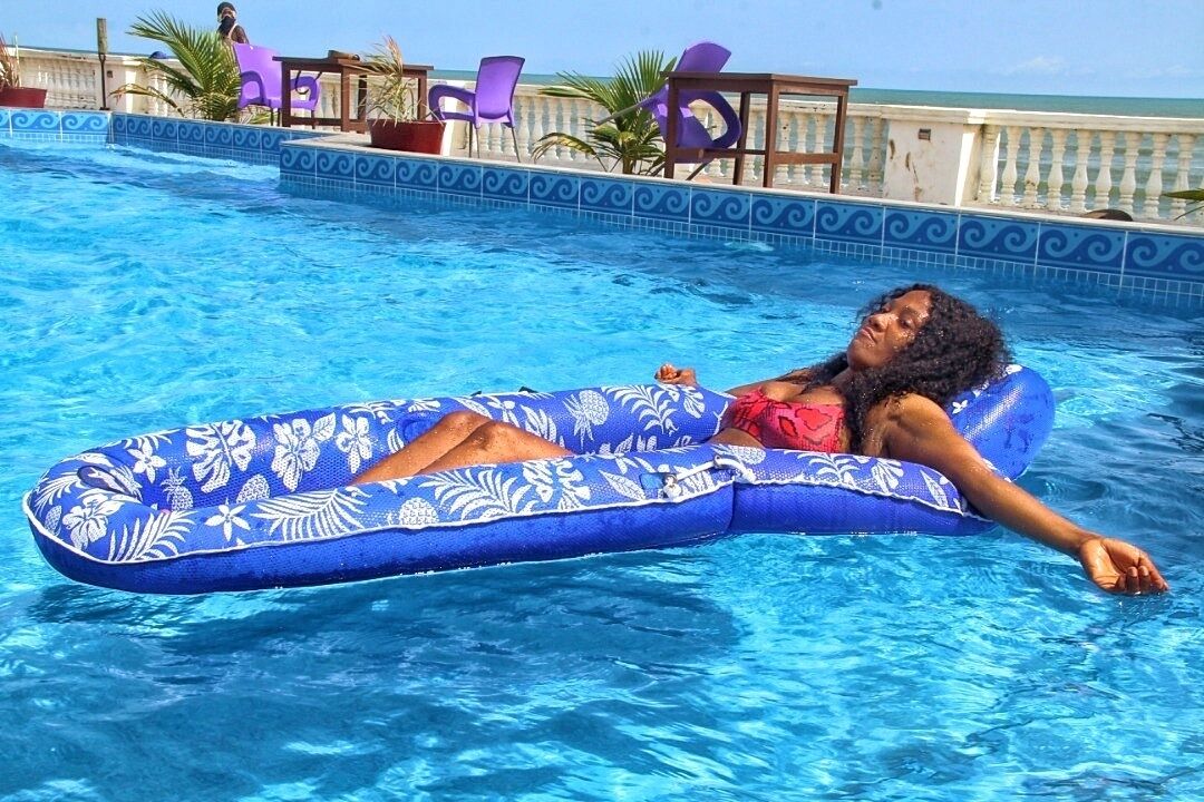 Una persona recostada sobre un flotador azul en una piscina con borde de azulejos y palmeras al fondo.