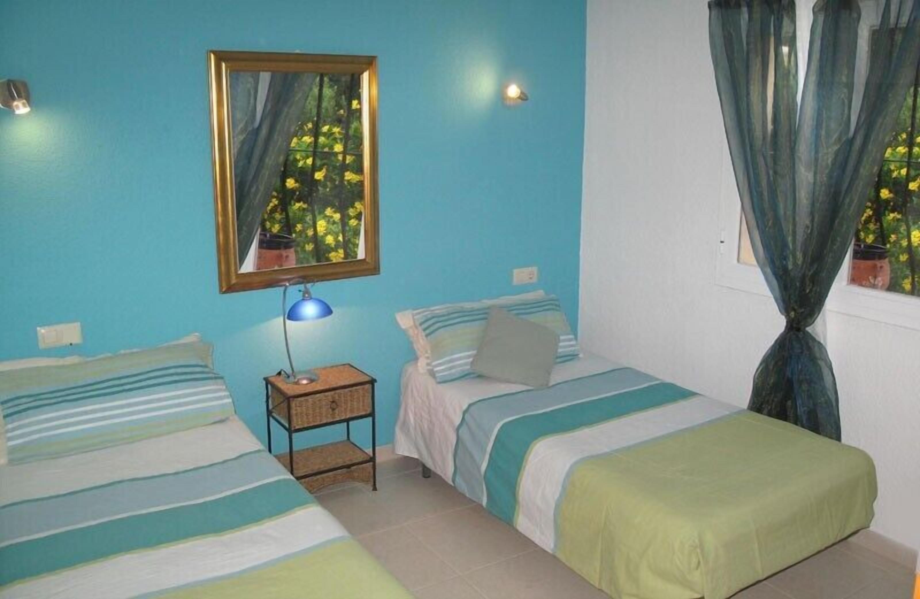 Una habitación con dos camas, una mesita de noche, una lámpara, un espejo y una ventana con cortinas.